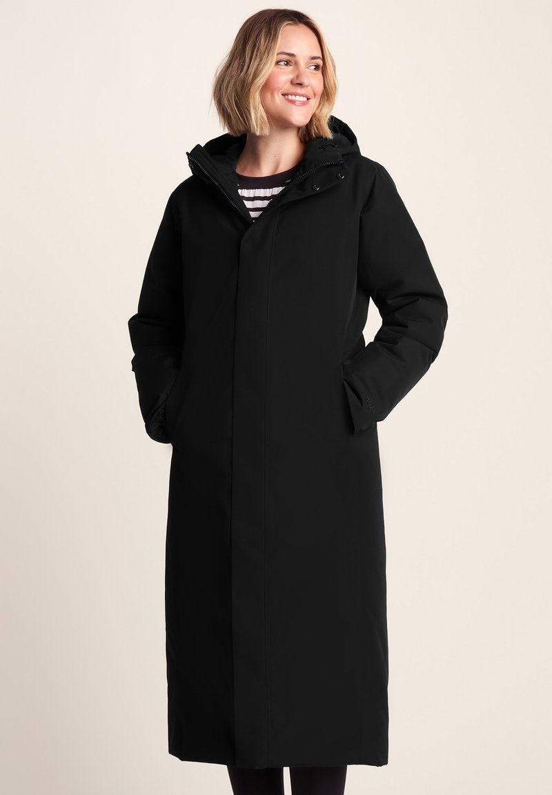 Woman smiling, wearing a long black hooded winter coat with hands in pockets, standing against a plain light background.