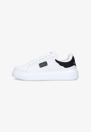 Sneaker bianca modello low-top con suola spessa, dettaglio nero sul tallone a forma di ali, piastra laterale in metallo e lacci bianchi su sfondo bianco.