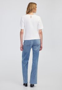 Weißes T-Shirt mit kurzen Ärmeln und Schlüsselaugen-Detail auf der Rückseite, kombiniert mit hellblauen, hochgeschnittenen Jeans mit fransigen Säumen und einer lässigen Passform.