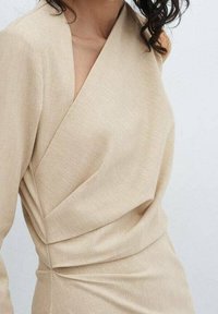 Beige, teksturoitu kietaisumekko syvällä V-aukolla ja epäsymmetrisillä sauman yksityiskohdilla. Vartalonmyötäinen siluetti ja laskokset vyötäröllä.