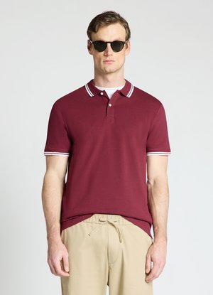 Uomo che indossa una polo rosso scuro con rifinitura bianca, pantaloni beige con coulisse e occhiali da sole neri, in piedi davanti a uno sfondo chiaro e uniforme.