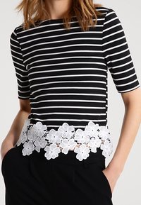 Mustassa ja valkoisessa raidallisessa t-paidassa on lyhyet hihat, ja siinä on valkoinen pitsikoristeinen helma. Pehmeä materiaali, vartalonmyötäinen muotoilu ja pyöreä pääntie.