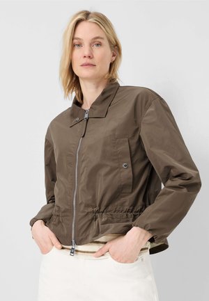 Frau mit kurzem blondem Haar, die eine braune Jacke mit Reißverschluss und Druckknopftaschen trägt, Hände in den Taschen, stehend vor einem schlichten hellen Hintergrund.