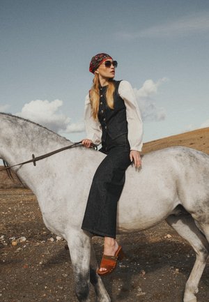 Donna con occhiali da sole, foulard, gilet e pantaloni a gamba larga seduta di lato su un cavallo bianco in un paesaggio aperto e arido.