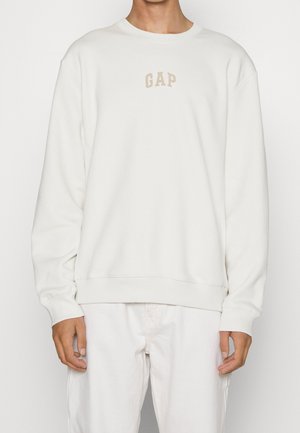 Person trägt ein weißes Sweatshirt mit Rundhalsausschnitt und beigem "GAP" Text auf der Brust sowie passende weiße Hose vor einfarbigem Hintergrund.