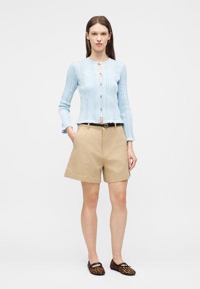 Γυναίκα που φοράει γαλάζια ζακέτα με κουμπιά, μπεζ σορτς ψηλόμεσα με μαύρη ζώνη και μπαλαρίνες με σχέδιο λεοπάρ, στέκεται με το ένα χέρι στην τσέπη.