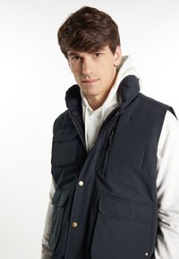 Gilet imbottito blu navy con colletto alto, due tasche frontali e tasca sul petto con zip. Presenta bottoni a pressione e una texture liscia.