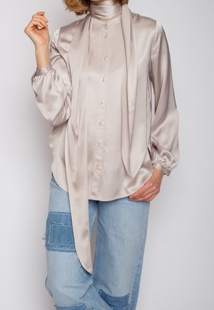 Mujer que lleva una blusa satinada beige brillante con lazos largos tipo bufanda combinada con vaqueros azul claro con grandes parches de denim.