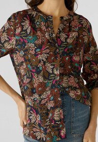 Femme portant une blouse boutonnée avec des motifs de feuilles multicolores en marron, rose, bleu sarcelle et magenta, associée à un jean en denim bleu.