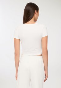 Donna con lunghi capelli castani, che indossa un crop top bianco aderente a maniche corte e pantaloni bianchi a vita alta, in piedi con la schiena rivolta.