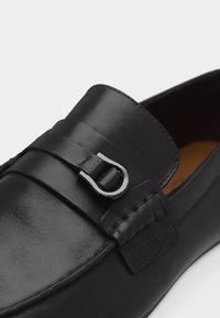 Mocassin en cuir noir avec détail de boucle en métal argenté sur le dessus, coutures visibles et finition lisse et polie.