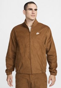 Brūnā kordura jaka ar augstu apkakli, priekšējo rāvējslēdzēju un elastīgām piedurknēm. Uz krūtīm ir balts Nike logo. Mīksta tekstūra.