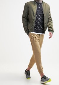 Chaqueta bomber verde oliva, sudadera negra con estampado, joggers beige y zapatillas negras/ grises con acentos en verde neón. Atuendo simple y casual.