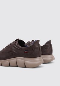 Zapatillas de cuero marrón con suela redondeada y gruesa. Presentan cordones y un pequeño parche de logo. Textura suave con detalles de costura.