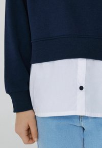 Marineblauer Sweatshirt mit gerippten Bündchen, über einem weißen Button-Down-Hemd mit dunkler Knopfdetaillierung getragen. Es sind helle blauen Jeans sichtbar.
