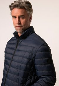 Námořnická modrá prošívaná bunda s horizontálním prošíváním, vysokým límcem, černým zipem a kulatou nášivkou na levém rukávu.