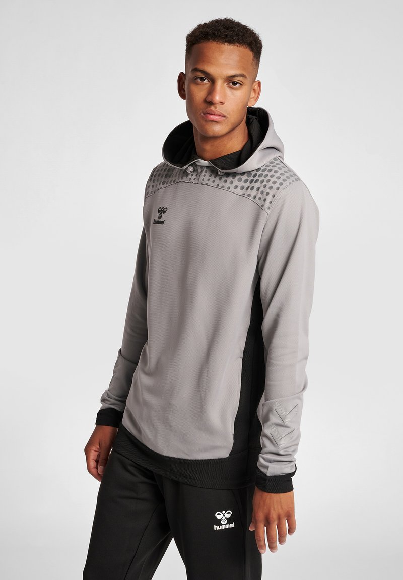 Giovane uomo che indossa una felpa con cappuccio hummel grigia e nera e pantaloni della tuta neri hummel, in piedi davanti a uno sfondo chiaro uniforme.