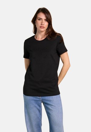 Donna con capelli lunghi e castani che indossa una t-shirt nera a maniche corte con testo in rilievo e jeans azzurri, in piedi con le mani dietro la schiena.
