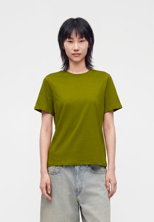Personne aux cheveux raides et foncés porte un T-shirt vert olive et un jean bleu clair, debout devant un fond blanc uni, face à l'avant.