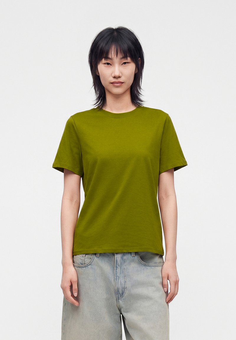 Personne aux cheveux raides et foncés porte un T-shirt vert olive et un jean bleu clair, debout devant un fond blanc uni, face à l'avant.
