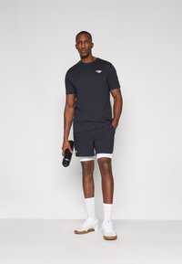 Black short-sleeve t-shirt with a logo on the chest, paired with black shorts featuring white accents, and white athletic shoes.