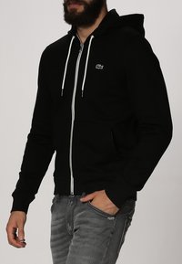 Zwarte rits hoodie van zacht materiaal, met een kangoeroezak, witte trekkoorden en een klein groen logo op de borst.