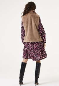 Kobieta ubrana w beżową kamizelkę z sztucznego futra, nosząc długą sukienkę w różowe, czarne i białe wzory z długim rękawem, w połączeniu z czarnymi, wysokimi kozaczkami na obcasie, stoi.