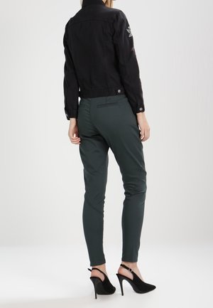 Vrouw draagt een zwart kort jasje, donkergroene strakke broek en zwarte hoge hakken slingback schoenen, staat met haar rug naar voren.
