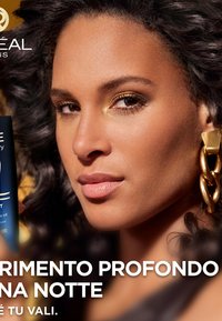 Una bottiglia blu scuro con un'etichetta che presenta un testo bianco, tenuta vicino a un volto con pelle abbronzata, capelli ricci scuri e orecchini dorati.