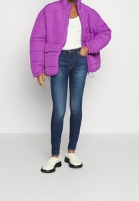 Chaqueta de plumas morada con costuras horizontales, camiseta sin mangas blanca, jeans ajustados azules y zapatos de lona crema con suelas negras.