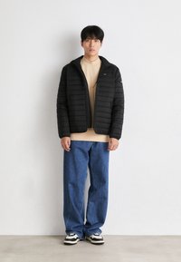Giubbotto imbottito nero con texture trapuntata, zip frontale e colletto alto. Abbinato a un top beige, jeans a gamba larga blu e sneakers nere.