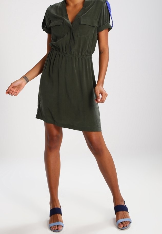 Femme portant une robe vert foncé à manches courtes avec une taille élastique et deux poches poitrine, associée à des talons ouverts bleu et marine.