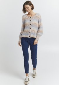 Stickad kofta i pastellrandigt mönster av blått, orange och krämfärgat, med rund halsringning, långa ärmar och stora mörka knappar. Matchad med blå jeans.