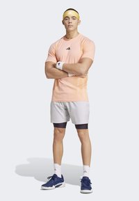Conjunto deportivo de Adidas que incluye una camiseta de manga corta color naranja claro, pantalones cortos grises, zapatillas azul marino y una cinta para la cabeza amarilla. Se incluyen muñequeras blancas.