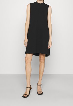 Femme portant une robe noire sans manches et des sandales noires à talons hauts, debout devant un fond clair uni.