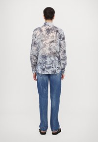 Camicia a fiori nei toni del blu e bianco, maniche lunghe, colletto con bottoni, indossata con jeans azzurri e calzature nere, vista da dietro.