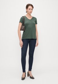 T-shirt verde a scollo a V, in tessuto leggero, abbinato a jeans skinny scuri e scarpe con tacco marroni. Piccola borsa grigia a tracolla.