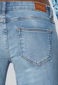 Svetlo modri denim kavbojki z zadnjim žepom, ki ima nastavljivo obliko in kontrastno šivanje; usnjen oznaka z napisom "ONLY" nad pasom.