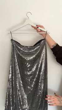 Abito con paillettes argento metallico su una gruccia bianca, caratterizzato da un design drappeggiato, texture liscia e spalline sottili regolabili.