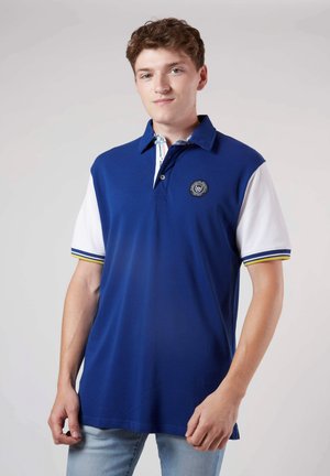 Polo bleu avec des manches blanches, arborant un logo sur la poitrine et des accents rayés sur le col et les bords des manches. Texture lisse.