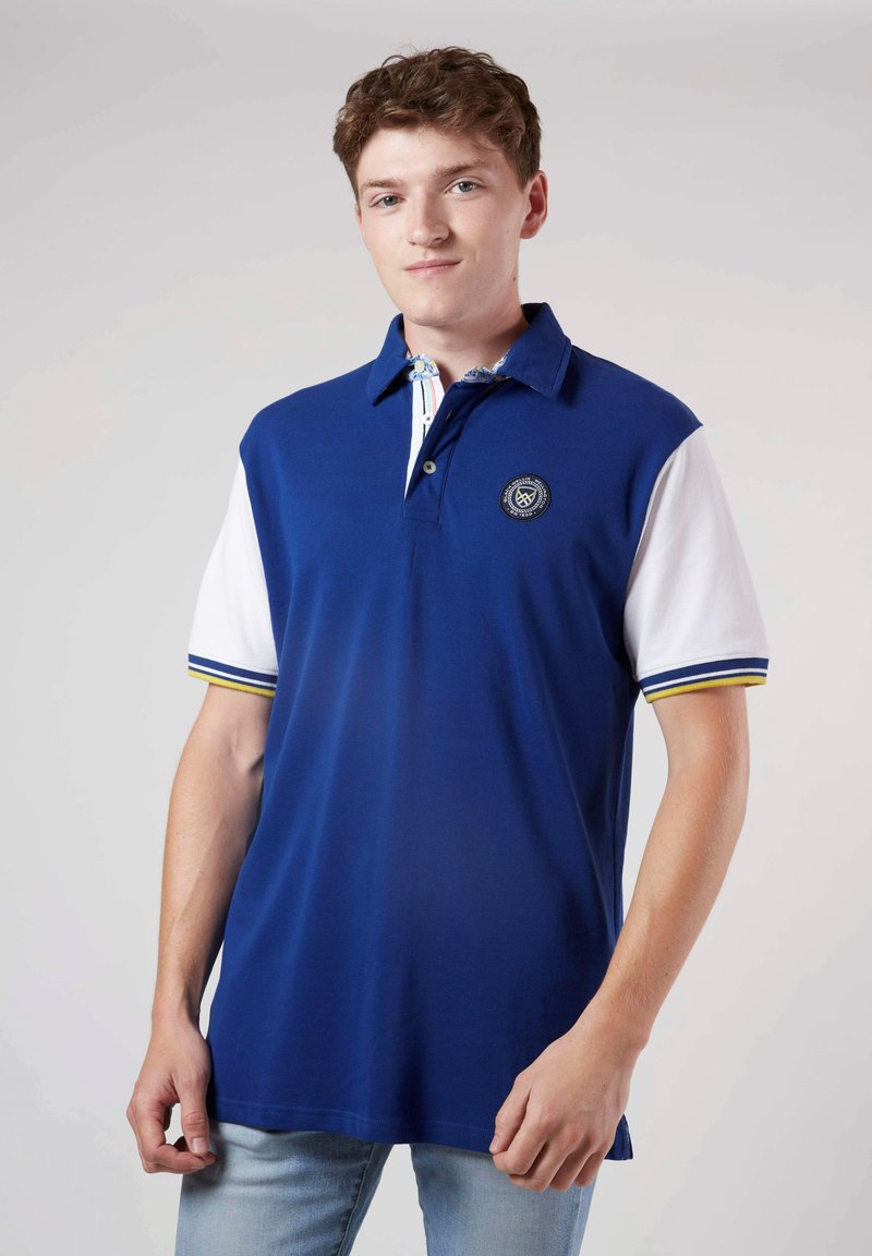 Polo bleu avec des manches blanches, arborant un logo sur la poitrine et des accents rayés sur le col et les bords des manches. Texture lisse.