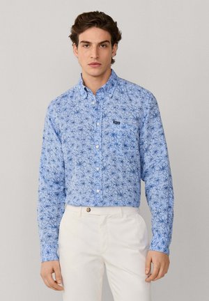 Joven con camisa azul de manga larga con botones y estampado floral, y pantalones blancos, posando frente a un fondo liso de color gris claro.
