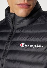 Juodas pūkuotas švarkas su aukštu kaklu ir matomomis siūlėmis. Yra baltas "Champion" logotipas su raudonu akcentu šalia užtrauktuko. Lygi tekstūra.