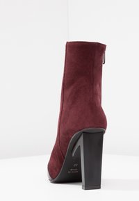 Bottine en daim bordeaux à finition texturée, talon bloc noir haut et détail de fermeture éclair latérale. Forme angulaire avec des bords lisses.