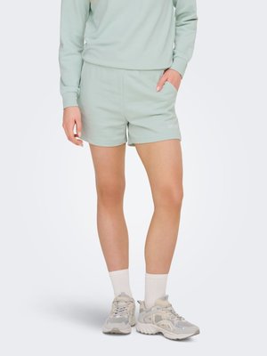 Person trägt hellgrüne Freizeitshorts, passendes Sweatshirt, weiße Socken und beige Sportschuhe, eine Hand in der Tasche, steht vor weißem Hintergrund.