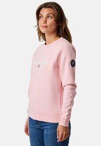 Felpa rosa con collo tondo, texture morbida, stampa bianca con la scritta "SEASIDE" e un badge sulla manica sinistra. Indossata con jeans blu.