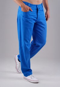 Blaue Baumwollhose mit lockerer Passform, ausgestattet mit einem Knopf- und Reißverschluss, Fronttaschen und getragen mit weißen Sneakern.