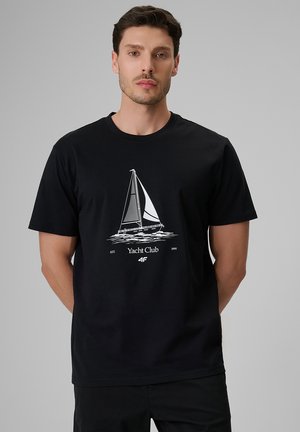 Mężczyzna w czarnej koszulce z białą grafiką żaglówki i napisem "Yacht Club" z przodu, stojący na jednolitym szarym tle.