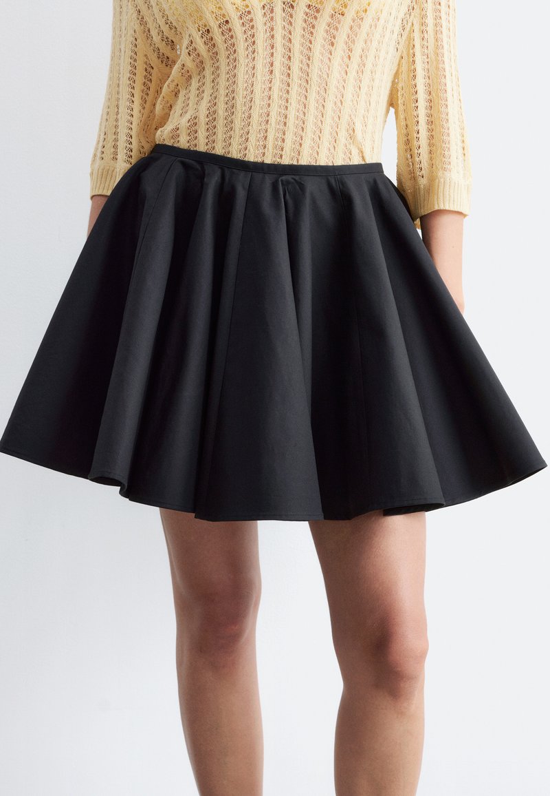 Femme portant une jupe plissée noire en ligne A et un haut tricoté jaune à manches trois-quarts, debout devant un fond clair.