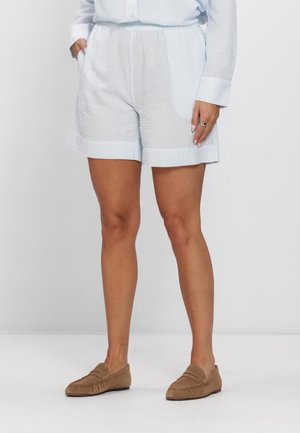 Person trägt weiße, strukturierte Shorts mit Taschen, ein passendes langärmeliges Oberteil und braune Slip-on-Loafer und steht vor einem schlichten Hintergrund.